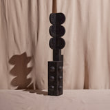 A wooden sculpture with a dark finish, consisting of several stacked circular forms on a vertical pole, set against a plain background.