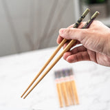 Hand holding a pair of wooden chopsticks with a blurred pack of chopsticks in the background.