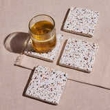 A set of four square terrazzo coasters with a multicolored pattern, placed on a beige fabric surface.