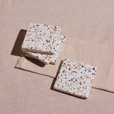 A set of four square terrazzo coasters with a multicolored pattern, placed on a beige fabric surface.
