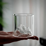 A hand holding a clear glass tumbler with a sculpted mountain design inside the base, set against a softly blurred indoor background.