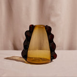 A conical amber glass vase with dark decorative elements placed against a pink textured background.
