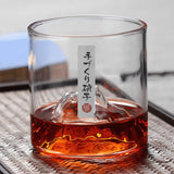 A clear glass tumbler with a mountain-shaped bottom inside, partially filled with amber-brown liquid, resting on a woven tray. A vertical white label with black Japanese characters and a small red seal is attached to the glass.