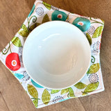 A white ceramic bowl placed on a square fabric coaster with a colorful floral and leaf pattern, photographed from above on a wooden surface.