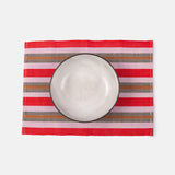 A handwoven placemat with red, pink, and brown stripes, holding a white dinner plate, placed on a white background.