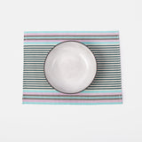 A handwoven placemat featuring a striped pattern in beige, pink, green, and blue, under a white dinner plate.