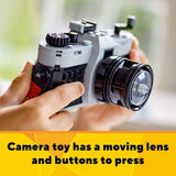 Toy camera with a moving lens and buttons held by a hand, on a blurred background.