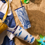 Two patterned tumblers on sand with a yellow bag and green toy nearby