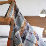 Person holding a plaid blanket over a bed with a wooden headboard.