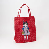 A red canvas tote bag with a repeating floral pattern and a central illustration of a black-and-white cat wearing a blue and pink dress, standing on a white background.