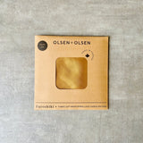 Olsen+Olsen Furoshiki fabric gift wrap in a square kraft paper envelope with a die-cut window showing the gold satin fabric inside, set on a textured gray background.