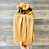 A hand holding a rectangular package wrapped in shiny mustard-gold paper with a large gathered bow on top, set against a rustic gray wooden plank background.