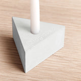 A light gray triangular candle holder base with a white cylindrical candle inserted into the top, set on a light wood grain surface.