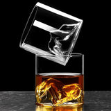 Two clear whiskey rocks glasses shown on a dark surface against a black background, one empty and one filled with amber liquor and a mountain formation.