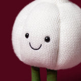 A close-up of a knitted garlic character toy with a white textured body, embroidered smiling face, and green stem-like legs against a dark red background.