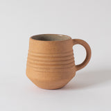 A beige ceramic mugs with a handle, placed on a neutral surface.