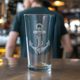 A clear drinking glass with an etched anchor design, standing on a wooden table with a blurred bartender in the background.