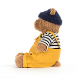 Teddy bear wearing a navy hat, striped shirt, and yellow overalls on a white background