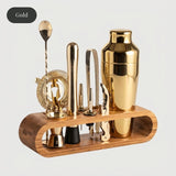 A gold cocktail bar tool set displayed on a curved wooden stand against a light gray background, with a black oval label in the top-left corner reading 'Gold'. The set includes a shiny gold shaker, strainer, bar spoons, muddler, and other bar accessories.