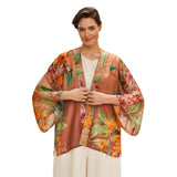 A woman modeling a lightweight open-front kimono jacket with wide sleeves, featuring a warm rust-brown base and a vibrant tropical floral print in orange, green, and pink, styled over a cream top and cream pants against a plain white background.