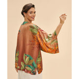 A woman wearing a loose, short-sleeve kimono-style jacket with a brown base and a tropical floral print in orange, green, and beige, photographed against a plain beige studio background.