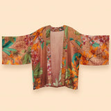 A lightweight open-front kimono-style jacket with wide sleeves, featuring a warm brown base with a vibrant tropical floral print in orange, green, and pink, displayed against a solid beige background.