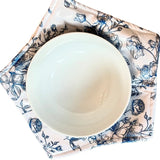 A white ceramic bowl placed on a square fabric napkin with a blue floral print on a light pink background, photographed from above on a white surface.