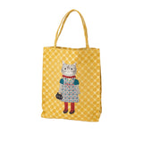 A yellow canvas tote bag with a repeating dot pattern and a central illustration of a gray cat wearing a colorful dress and holding a black purse, shown on a plain white background.