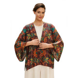 A woman wearing a lightweight open-front kimono-style top with a multicolor abstract print in brown, orange, teal, and green over a white top and cream pants, photographed against a plain white studio background.