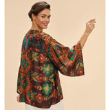 A woman wearing a loose, short-sleeved kimono jacket with a colorful geometric and floral pattern in brown, orange, teal, and gold, shown from a three-quarter back view against a solid beige background.