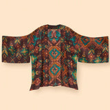A short-sleeved open-front kimono jacket featuring a complex geometric and tribal-inspired pattern in brown, teal, orange, and gold, displayed against a solid beige background.