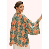 A woman wearing a loose, patterned kimono jacket with wide sleeves, photographed against a plain light pink background. The jacket has an allover geometric medallion print in teal green, orange, and cream.