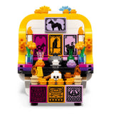 A LEGO Day of the Dead (Dia de los Muertos) ofrenda altar set, featuring a yellow and white structure decorated with purple and black animal tiles, orange flower bricks, candles, and a skull at the base, on a solid white background.