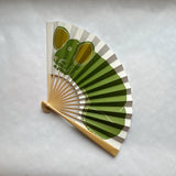 A traditional folding hand fan with a light wood handle, featuring a printed green rat illustration, displayed on a light gray textured background.