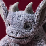 A close-up of a gray furry gargoyle toy with pointed ears, dark eyes, and a stitched smile against a dark red background.