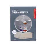 Kikkerland Galileo Thermometer in its retail packaging, featuring a 4-inch glass sphere with three colored liquid thermometers inside, resting on a wooden base.