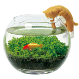 Glass fishbowl with a goldfish, green plants, and a cat figurine on the rim.
