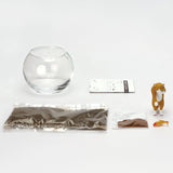 A DIY cat figurine kit laid out on a white background, including a clear glass bowl, a small brown-and-white cat figure, a packet of dark gravel, a packet of brown seeds, and a small orange fish.