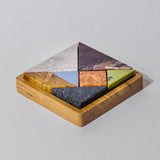 A colorful gemstone tangram puzzle with seven pieces, some in shades of red, orange, yellow, green, blue, and gray, displayed on a wooden board.