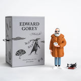 Collectible art toy figure of a man in an orange coat with a box featuring Edward Gorey branding.