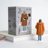 A gray illustrated product box stands on stacked books beside a small figurine wearing an orange-brown coat and blue pants, against a clean white background.