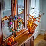 A Halloween-themed decorative wreath with colorful pumpkins, bats, skulls, and spiderweb details hangs on a wooden cabinet door. On the cabinet sill, a translucent amber glass vase holds dried orange autumn leaves, with small orange pumpkins resting on a book in the foreground.