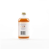 A clear glass bottle filled with amber liquid, shown from the back against a plain white background. The bottle has a white label with a barcode and small instructional text, and a gold screw cap.