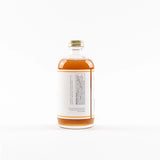 A clear glass bottle filled with amber liquid, featuring a white label with black text and a gold screw cap, shown against a plain white background.