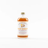 A bottle of Wood Stove Products Hot Toddy Honey, Lemon & Ginger syrup, filled with amber liquid and featuring a white label with orange text, set against a plain white background.
