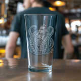 A clear glass pint glass with a white etched design of a sailing ship and octopus, standing on a wooden bar counter with a blurred bartender in the background.