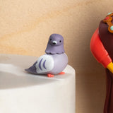 A cute, stylized toy pigeon figurine in shades of lavender purple, white, and bright orange feet, set against a clean light gray background.