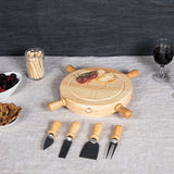 A wooden charcuterie board set with four matching utensils, arranged on a light gray tablecloth with food props including fruit, crackers, and a wine glass in the background.