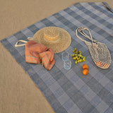 Blue checkered picnic blanket with a straw hat, orange, fruits, and a bag on a beige surface