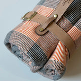 Folded picnic blanket with plaid pattern and brown leather straps on a light gray background